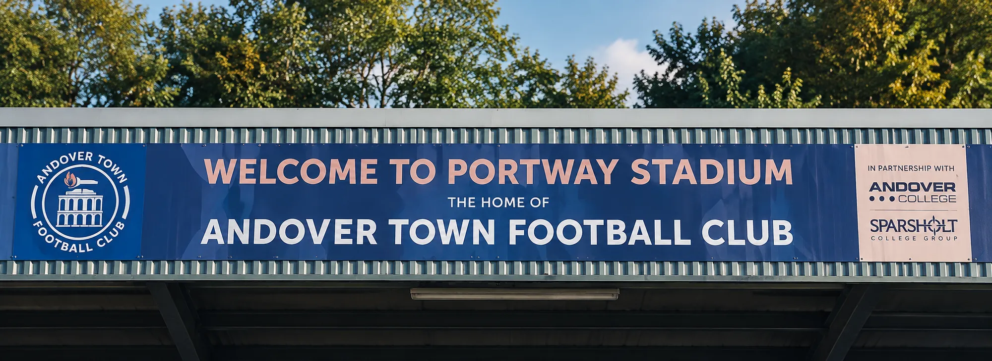 The main stand roof at Portway Stadium under trees, with a banner reading ‘Welcome to Portway Stadium, the home of Andover Town Football Club’ beside the Club crest, and Andover College and Sparsholt College Group named as partners.