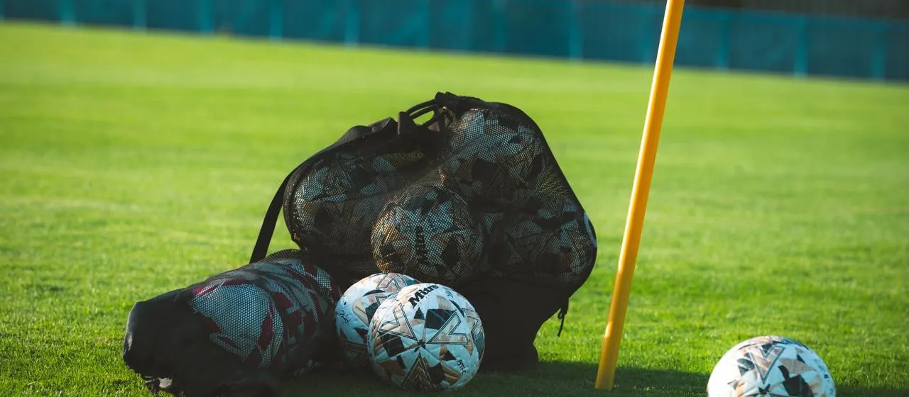 A ball bag and match balls on the grass beside a yellow corner flag, with the blue perimeter fence behind.